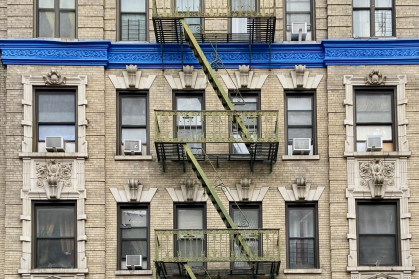 The facade of a residential prewar brick building with a green fire escape and a bright blue cornice in West Harlem, Manhattan, New York City